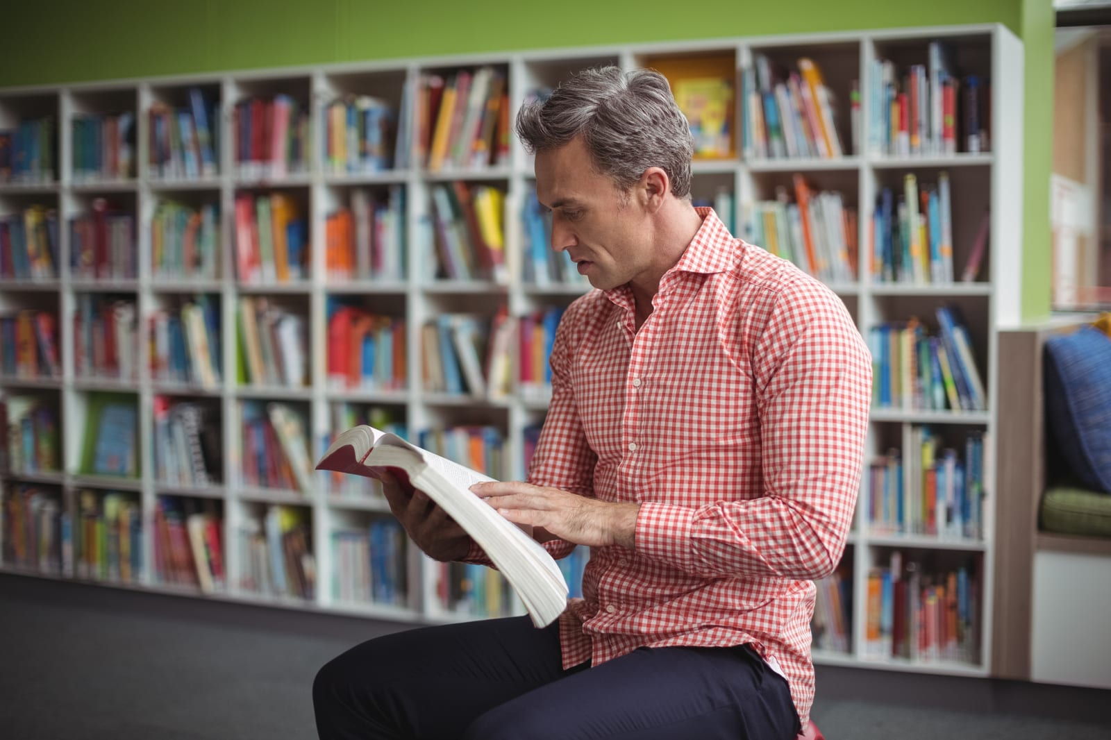 Homem sentado numa área de leitura de livraria, folheando um livro de capa dura perto de uma estante.