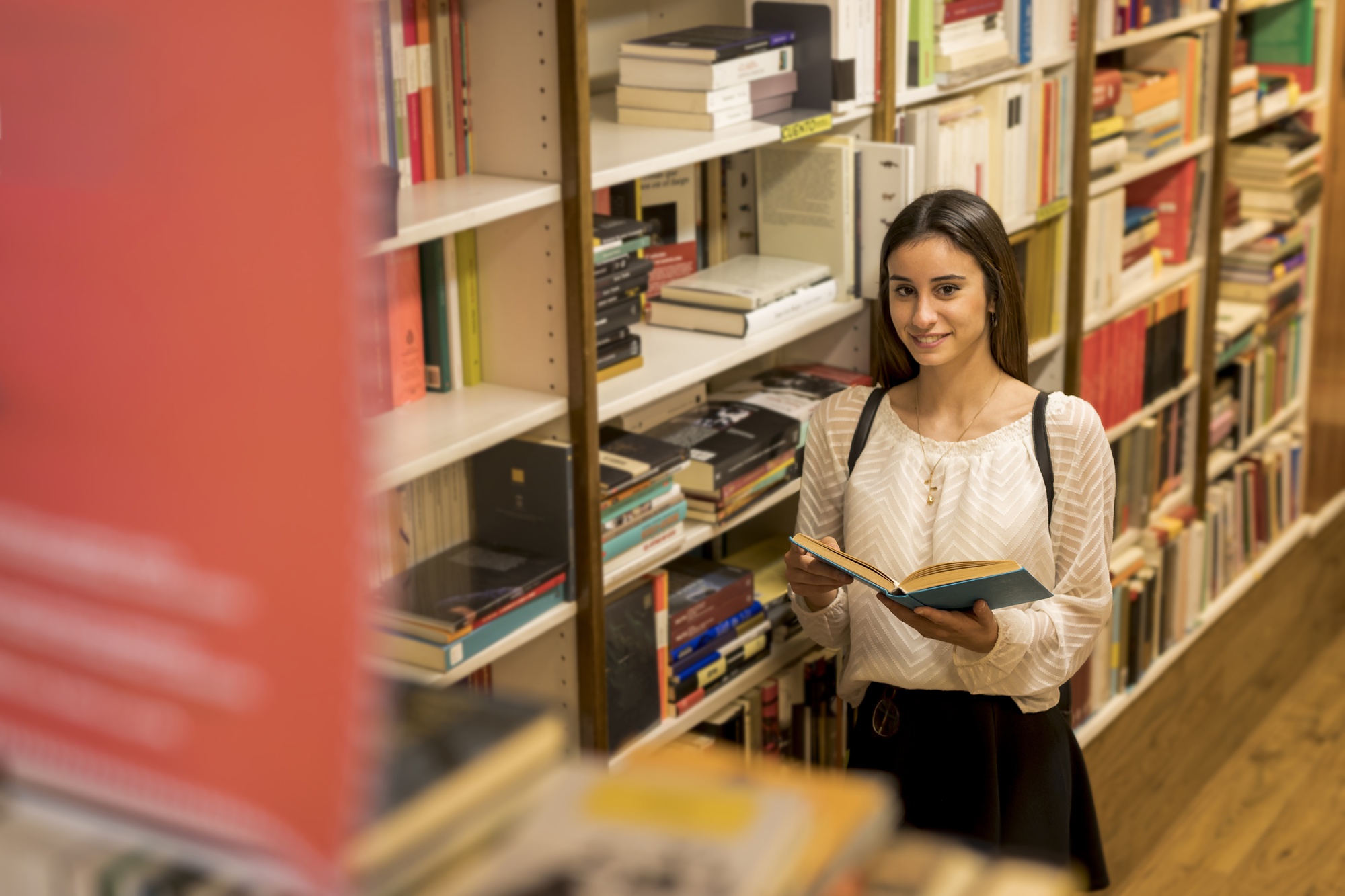 Mulher folheando um livro de capa azul entre estantes repletas de livros numa livraria.