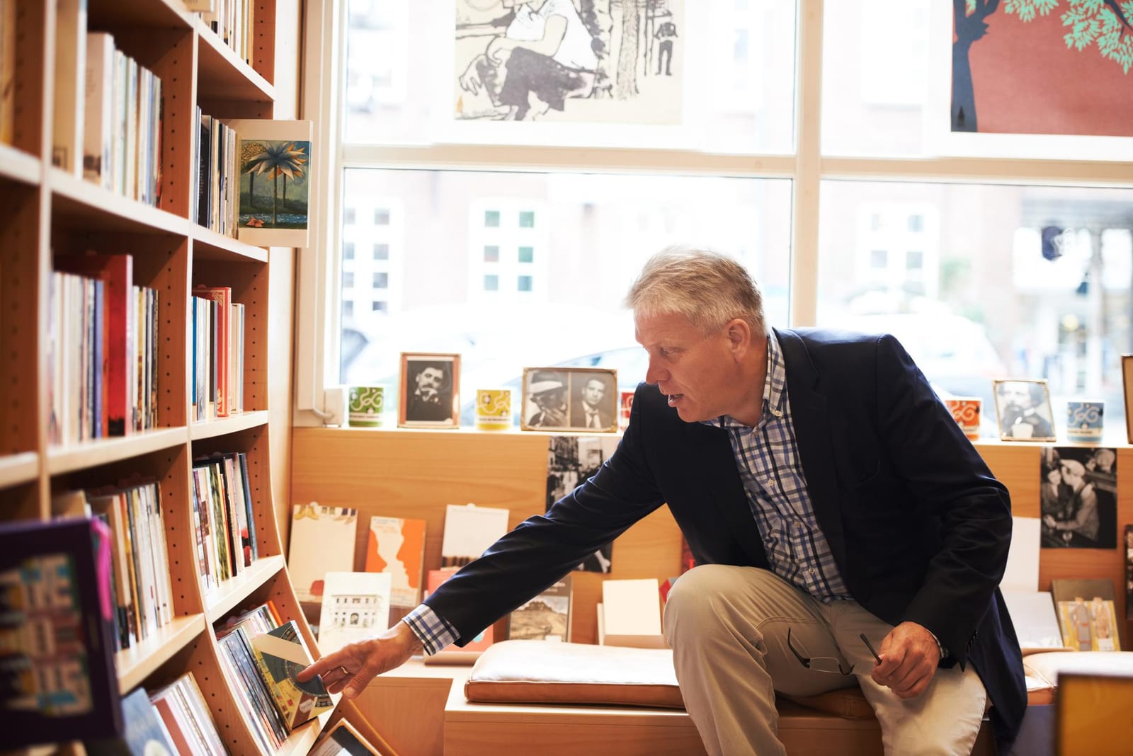 Homem em pé numa livraria, esticando o braço para tirar um livro da prateleira perto da vitrine.
