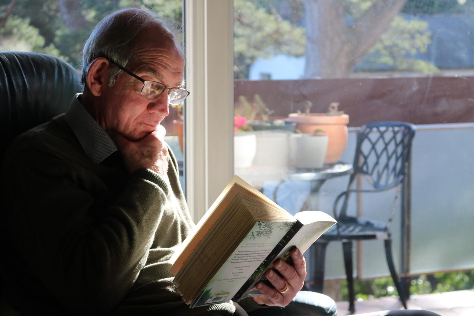 Homem mais velho, sentado perto de uma janela, concentrado na leitura de um livro grosso de capa dura.