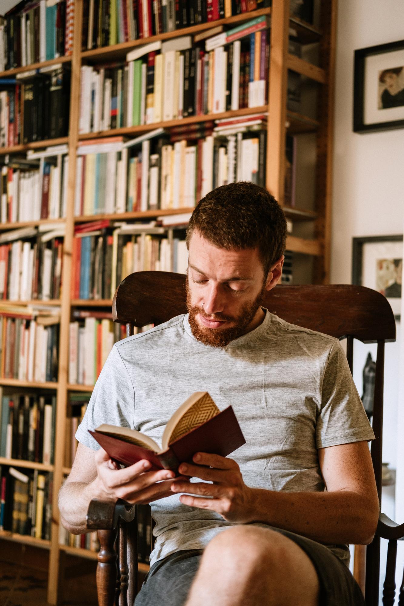 Homem lendo um livro numa poltrona de couro, com uma estante de livros desfocada ao fundo.